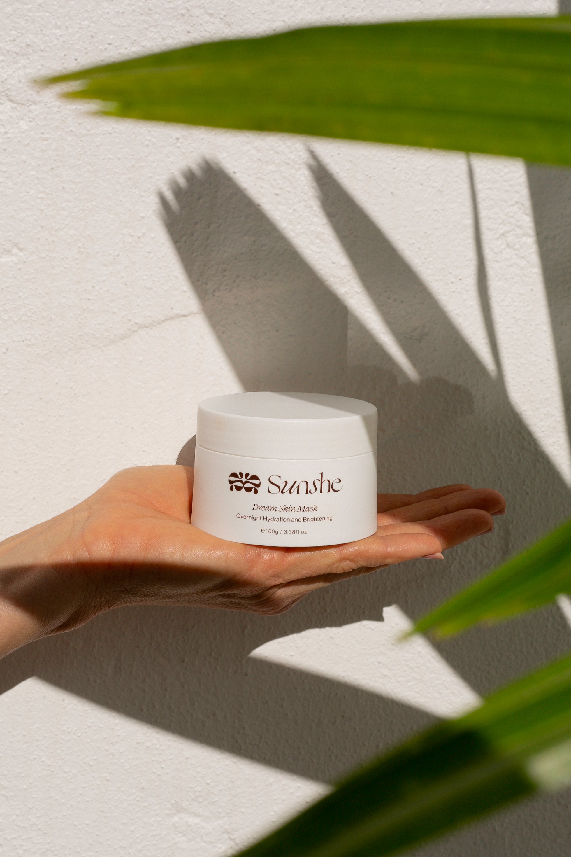 Hand holding a white jar labeled 'Sculpe' against a light background with palm leaves.