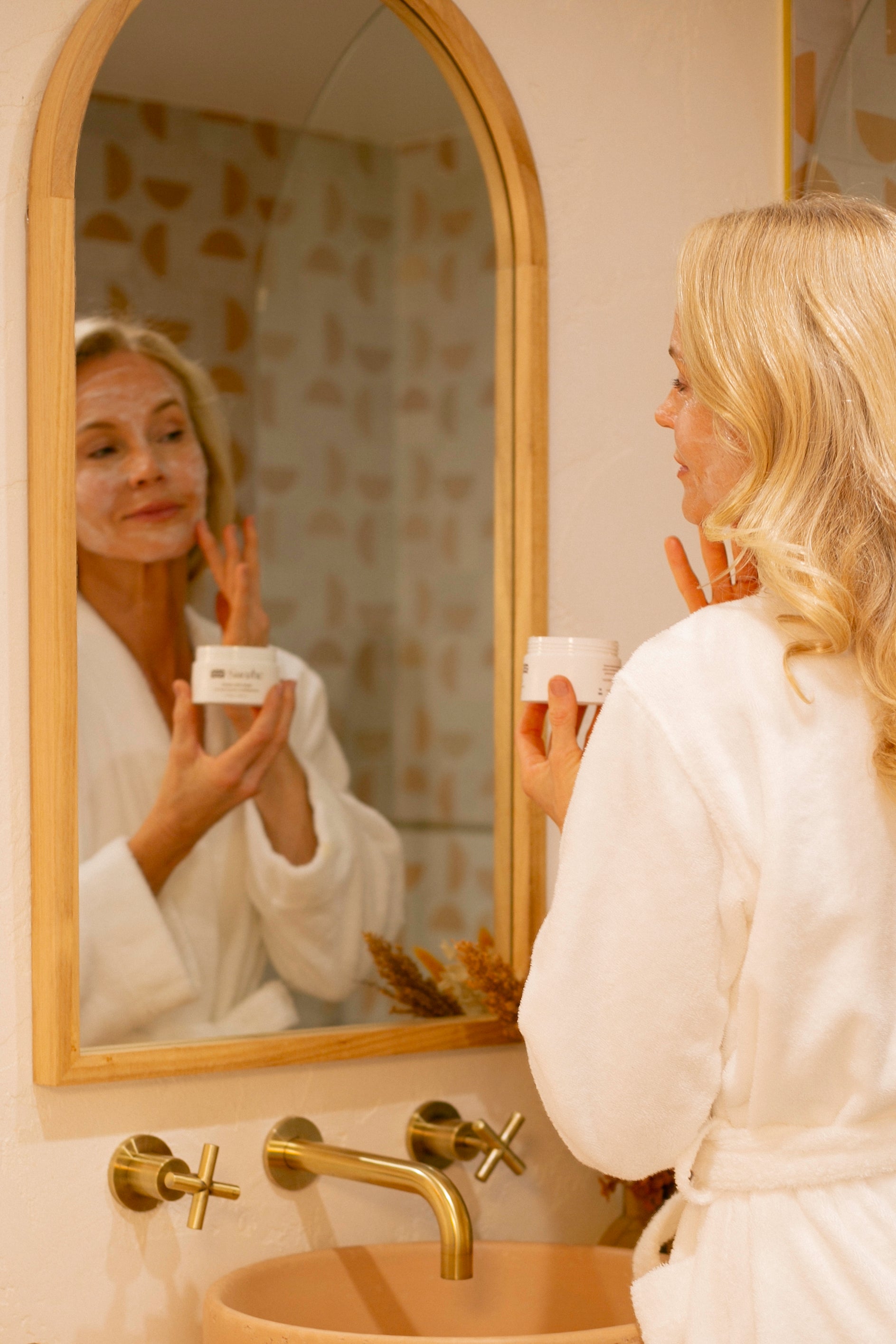 Woman in a white robe applying cream in front of a mirror with gold-framed mirror and brass faucet.
